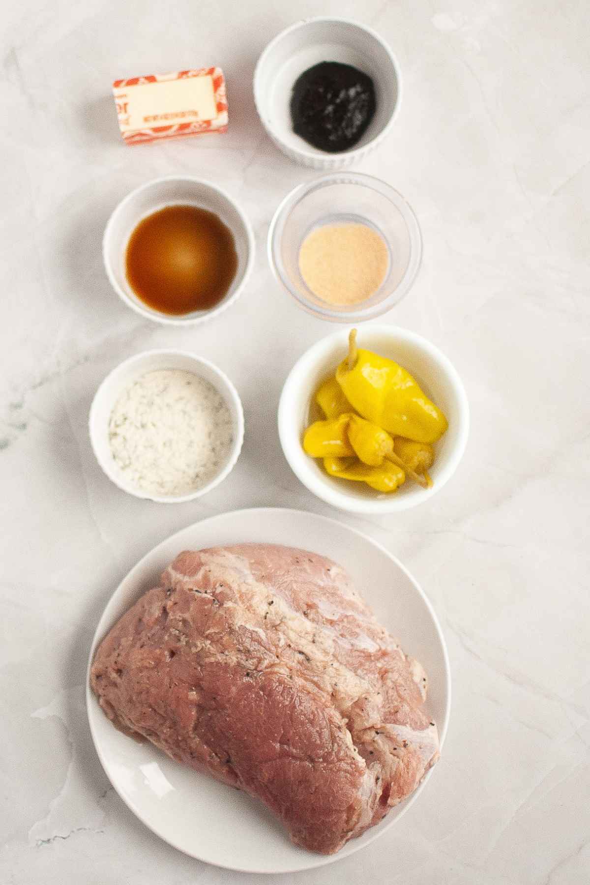 Overhead view of ingredients including a raw pork roast, pepperoncini peppers, butter, seasonings, and sauces in small bowls.