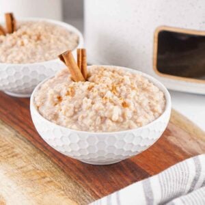 Two white bowls filled with creamy rice pudding on a wooden board, each topped with cinnamon sticks and a sprinkle of cinnamon.