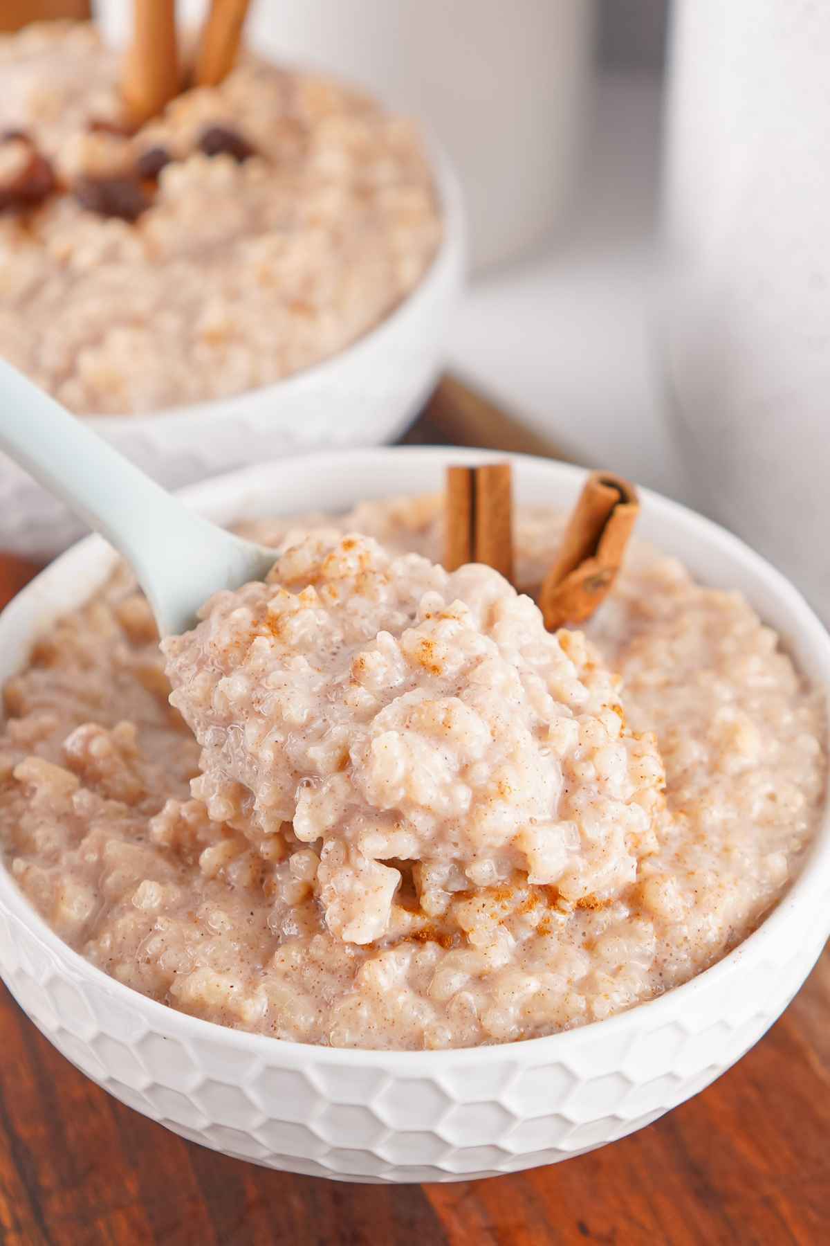A close-up of creamy rice pudding being scooped from a white textured bowl with a spoon, garnished with cinnamon sticks.
