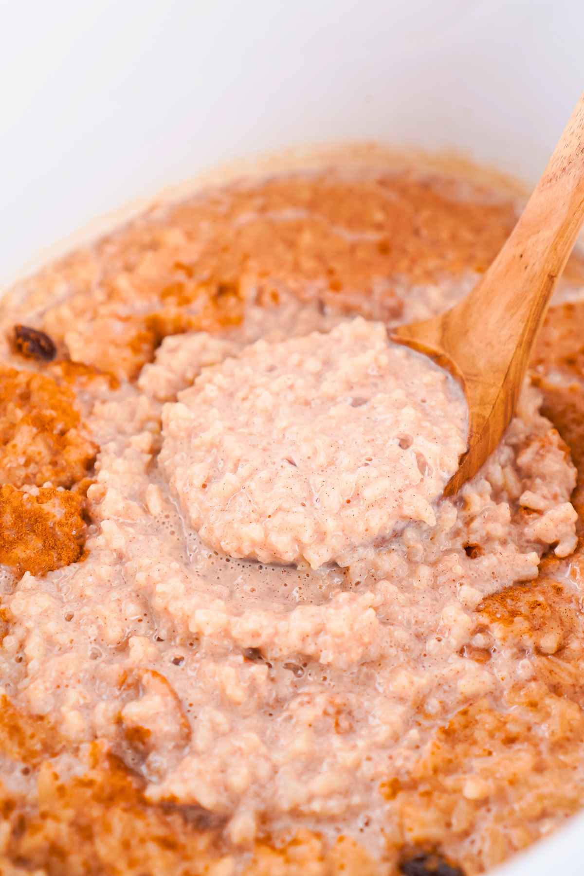A wooden spoon stirring creamy rice pudding inside a slow cooker, showing a thick and textured consistency with bits of cinnamon.