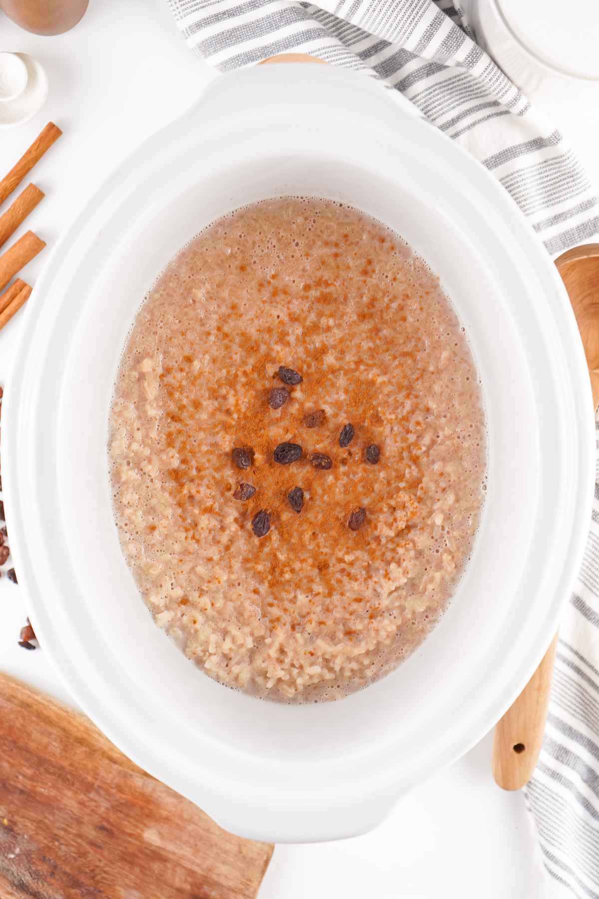 Overhead view of rice pudding in a white slow cooker sprinkled with cinnamon and raisins, surrounded by wooden utensils and a striped towel.