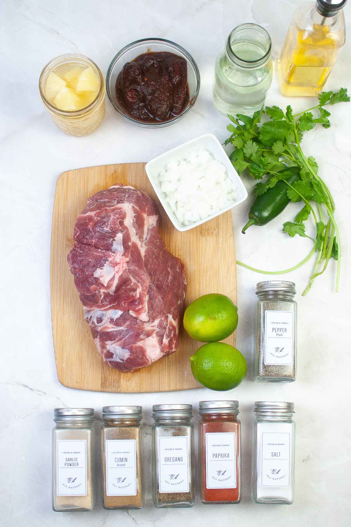 Raw pork shoulder on a cutting board surrounded by fresh cilantro, jalapeno, limes, diced onion, pineapple, chipotle peppers, spices, vinegar and oil.