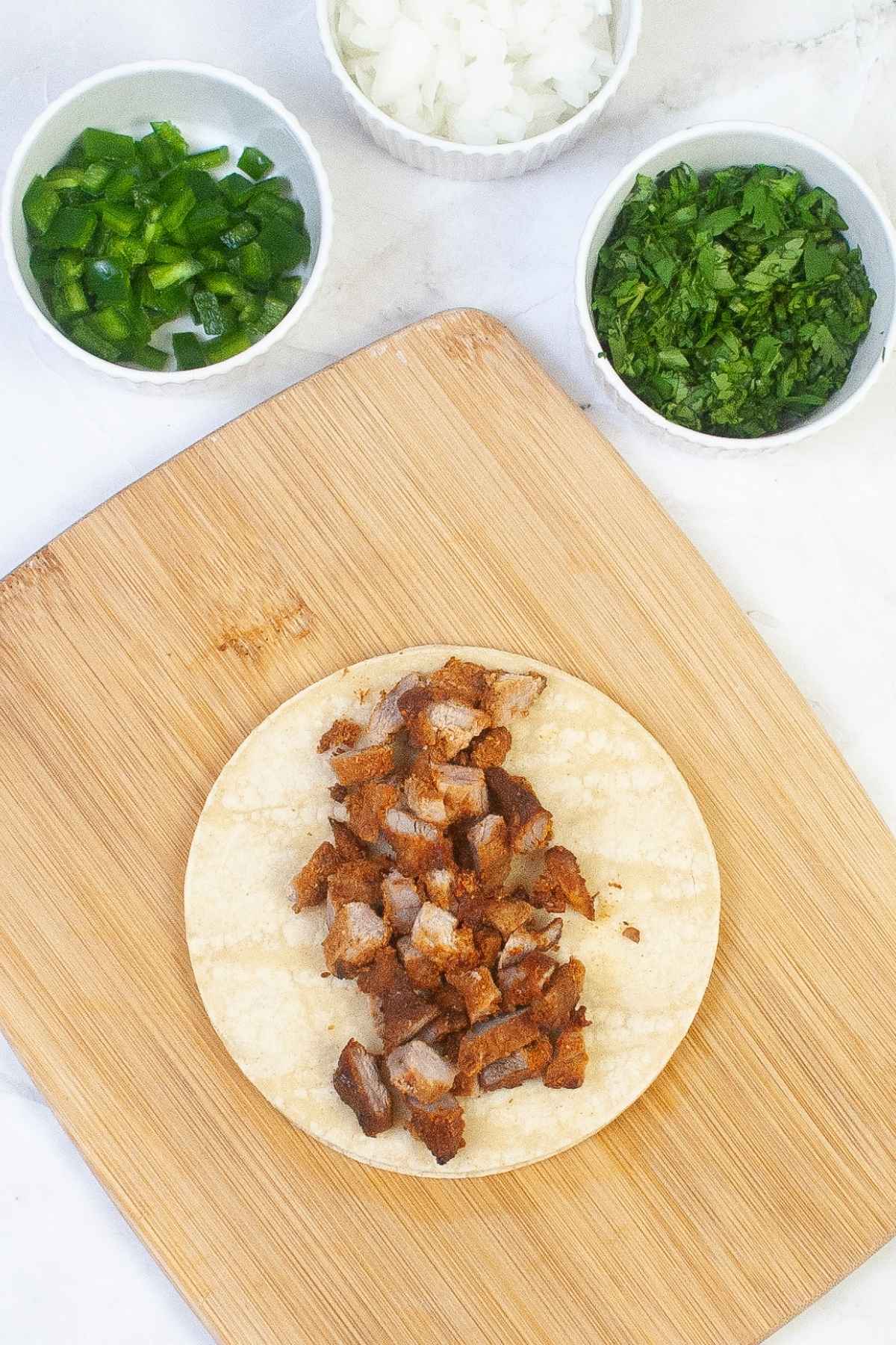 Corn tortilla on a wooden board filled with chopped pork tacos al pastor with small bowls of diced onion, jalapeno, and cilantro.