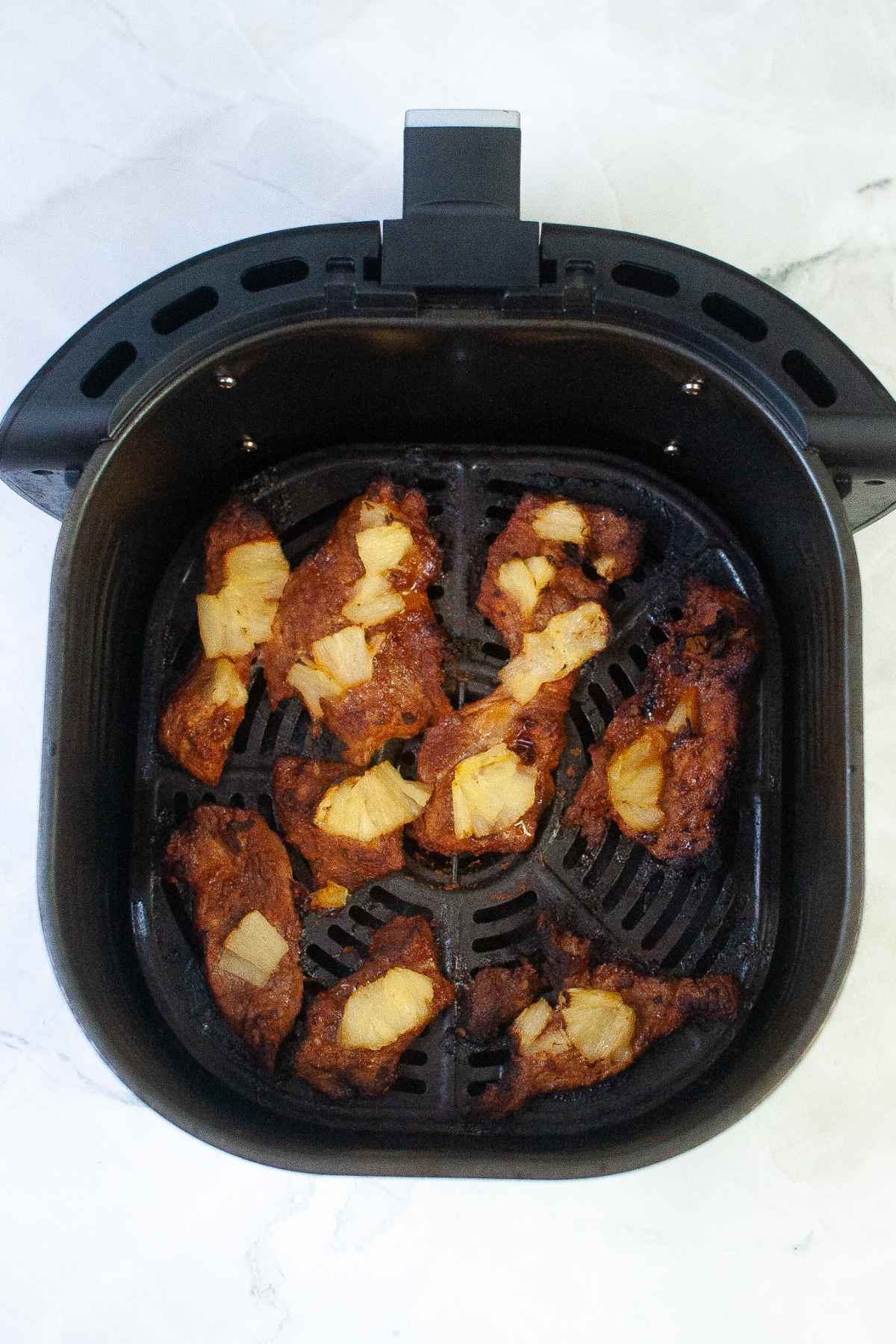 Slices of air fried pork with pineapple chunks cooked in an air fryer basket.