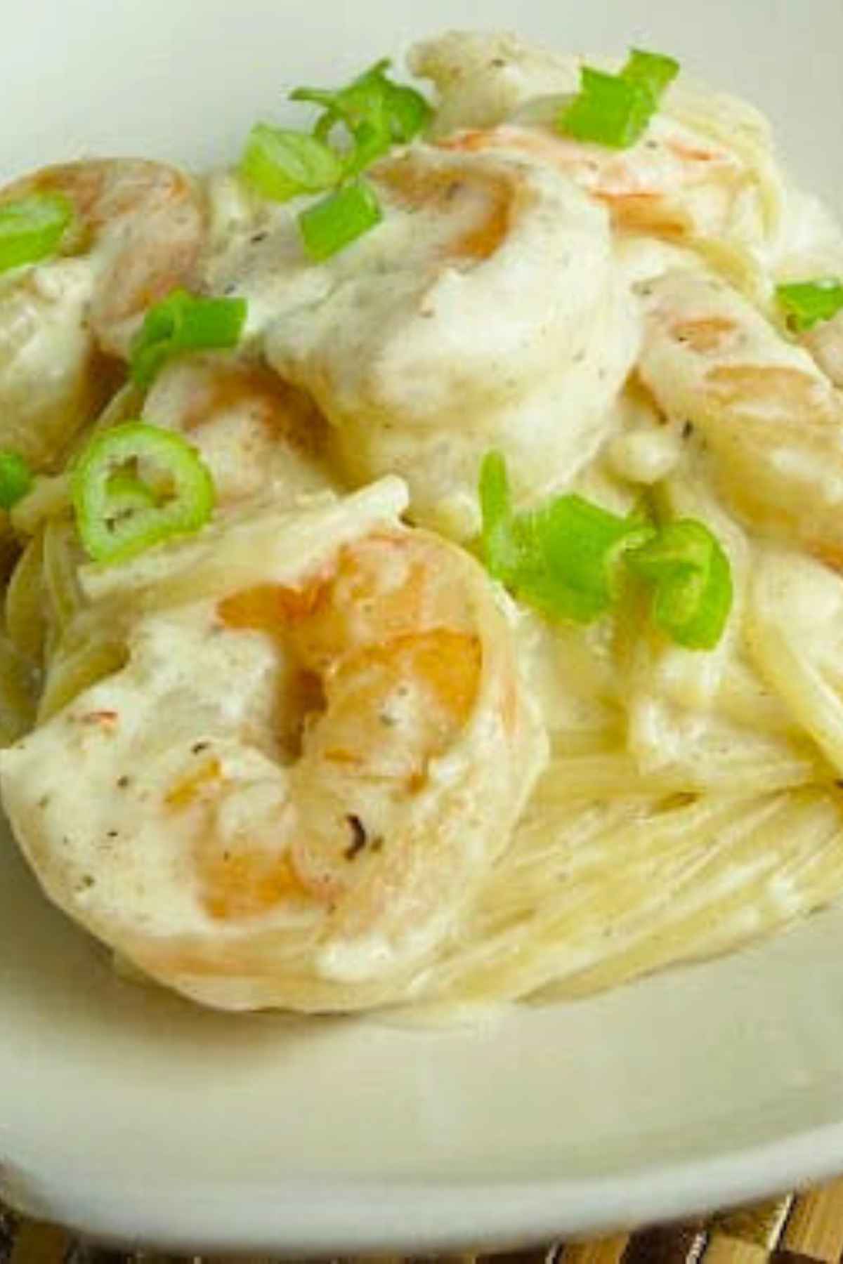 A plate of creamy shrimp linguine topped with sliced green onions, featuring large shrimp coated in a rich white sauce over thin pasta.