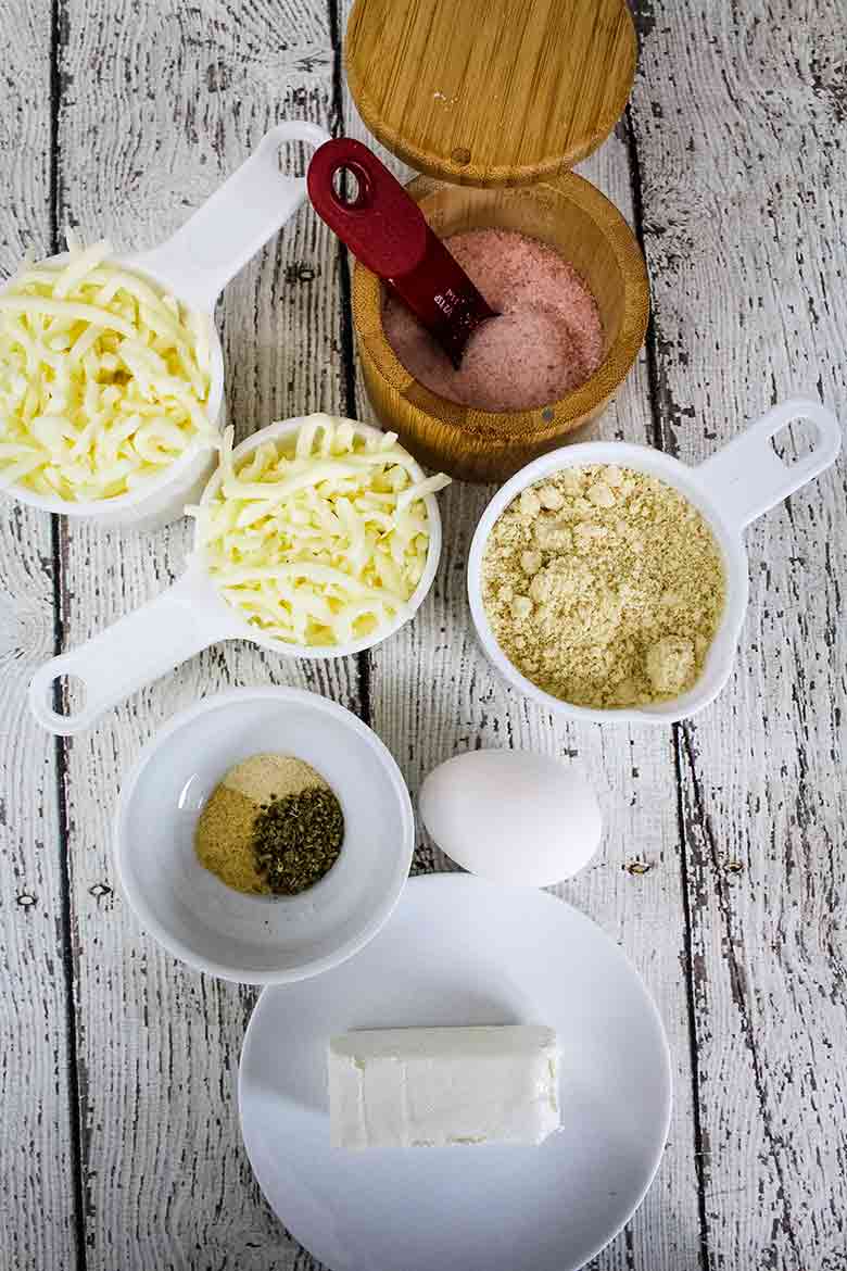 Ingredients for mozzarella pizza crust including shredded cheese, almond flour, cream cheese, egg, seasonings, and pink salt arranged on a wooden background.