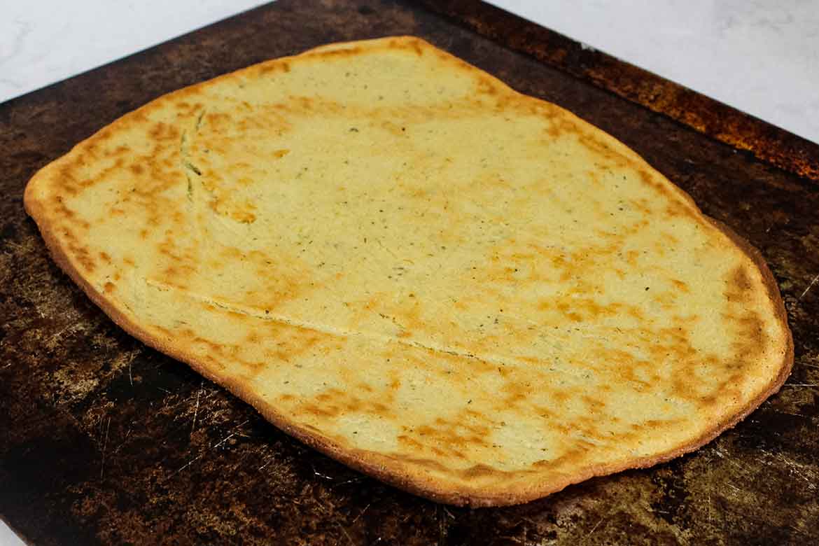 Baked keto pizza crust on a dark baking sheet, golden brown and slightly crisp around the edges.