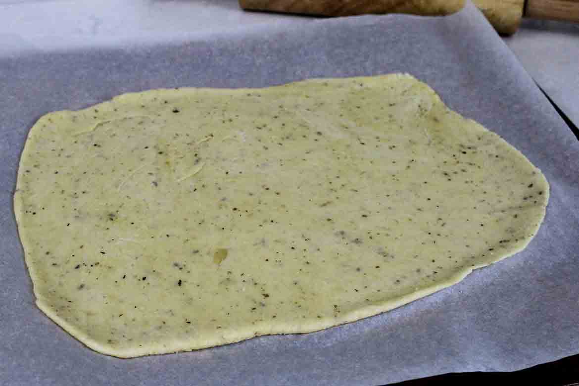 Rolled out keto pizza dough on parchment paper, evenly spread and speckled witih herbs, prepared for baking.