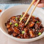 A bowl of General Tso Chicken topped with sesame seeds and sliced green onions, with chopsticks picking up a piece.