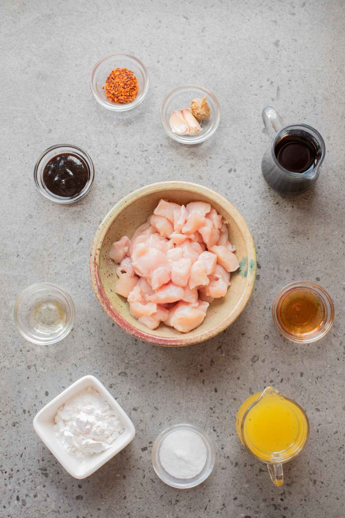 A flat lay of ingredients including raw diced chicken, sauces, spices, cornstarch, and broth arranged around a bowl.