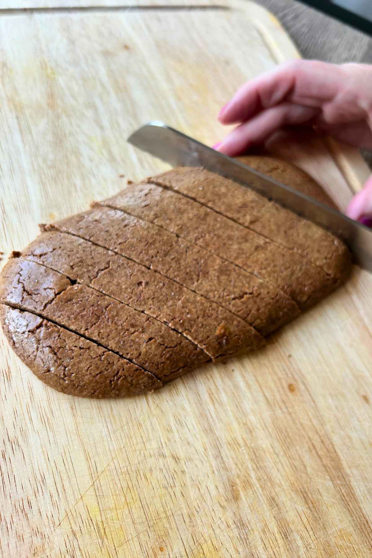 A hand slicing freshly baked biscotti dough into strips on a wooden cutting board.