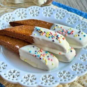 Finished Baileys biscotti half-dipped in white chocolate and topped with colorful round sprinkles, arranged neatly on a white lace-patterned plate.