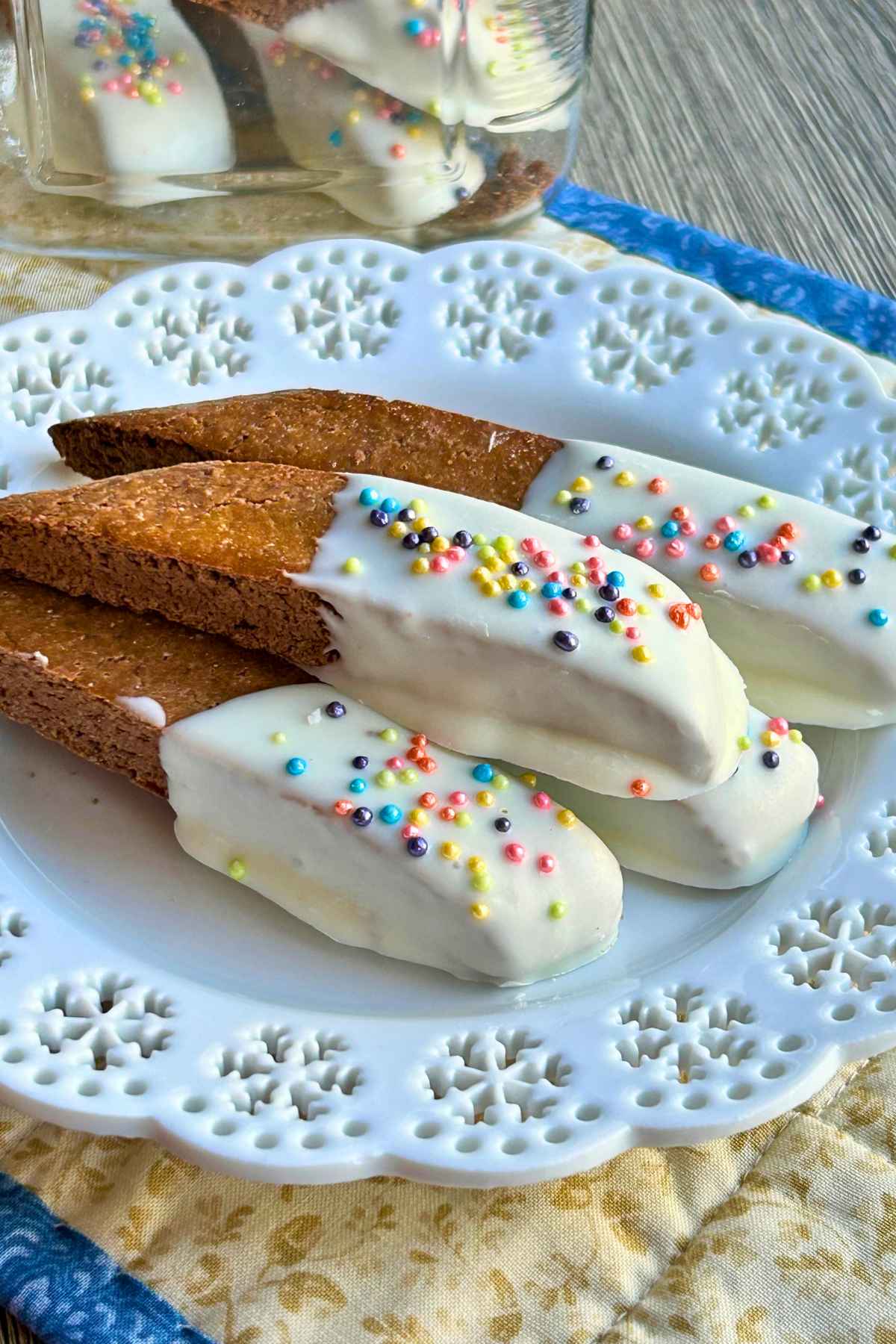 Finished Baileys biscotti half-dipped in white chocolate and topped with colorful round sprinkles, arranged neatly on a white lace-patterned plate.