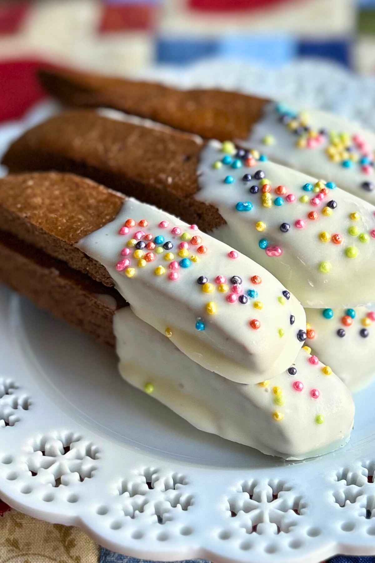 Finished Baileys biscotti half-dipped in white chocolate and topped with colorful round sprinkles, arranged neatly on a white lace-patterned plate.
