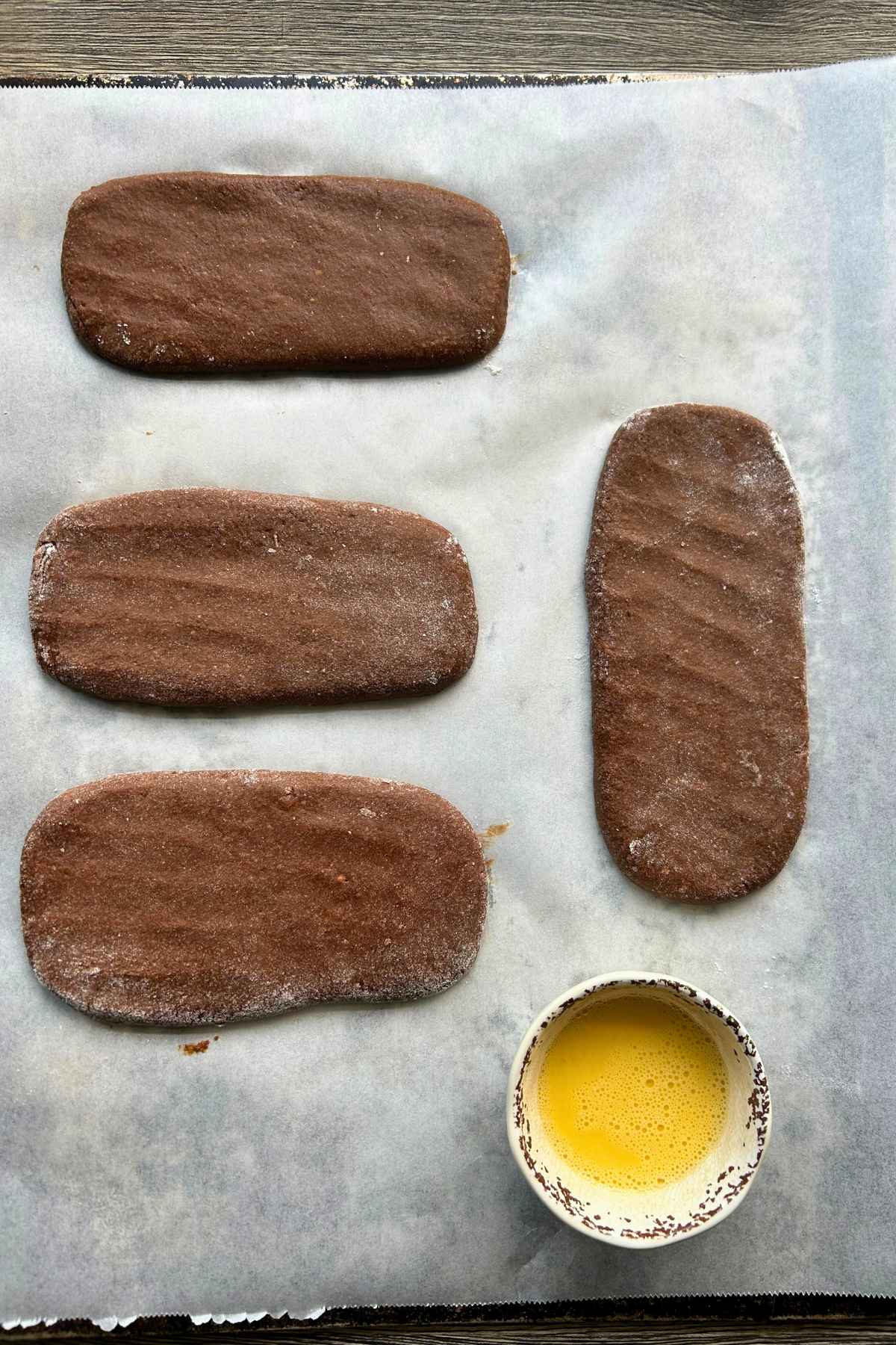Four unbaked chocolate colored dough logs on a parchment lined baking sheet beside a small bowl of whisked egg wash.