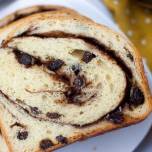 A thick slice of cinnamon raisin bread on a white plate, showing a detailed swirl of cinnamon and pockets of raisins in the soft crumb.