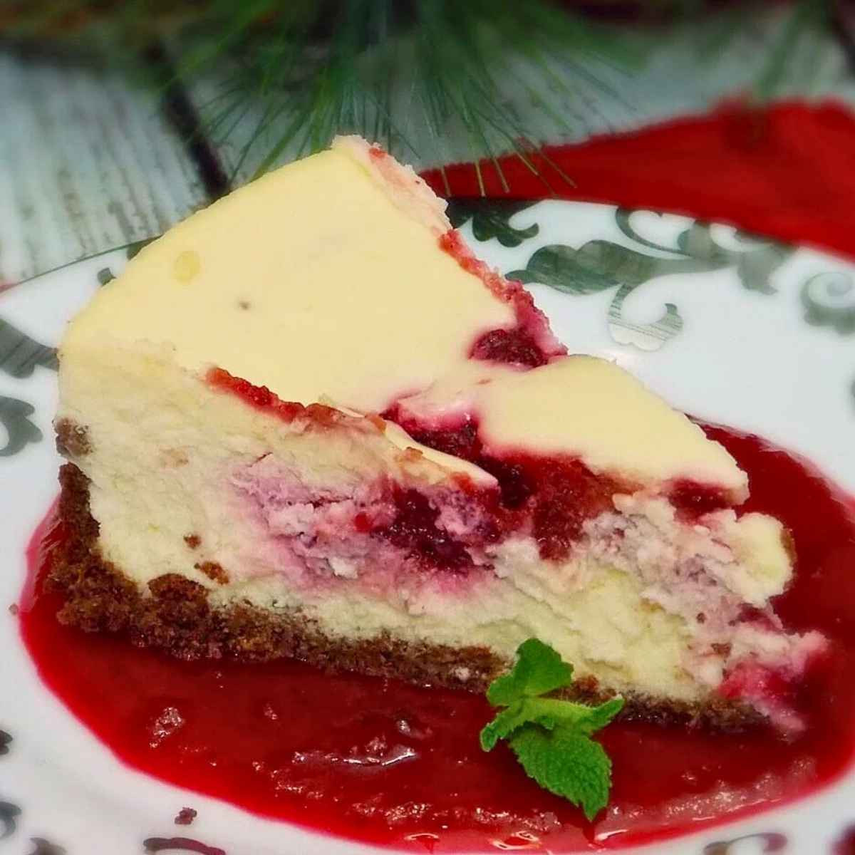 One slice of Cranberry Swirl Cheesecake sitting on a pool of blended amaretto cranberry sauce garnished with fresh mint on a silver trimmed dessert plate
