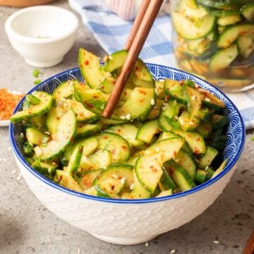 A white bowl with blue design filled with cucumber kimchi with a pair of chopsticks picking up some pieces of cucumber.