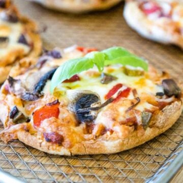 English Muffin Pizzas on an air fryer tray topped with mushrooms, red peppers, onions, and a fresh basil sprig.