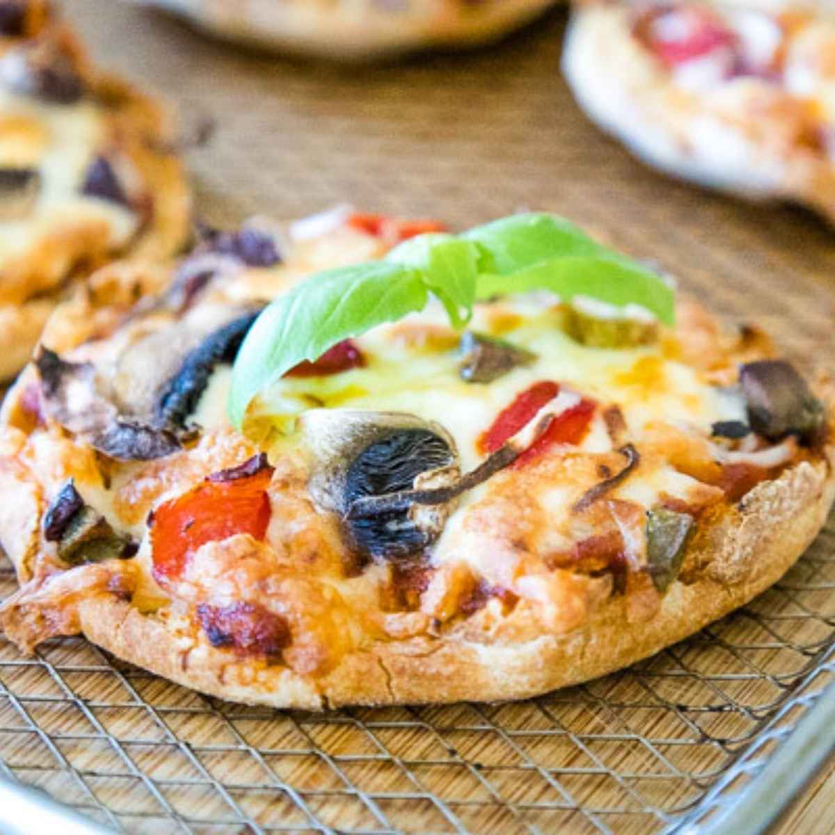 English Muffin Pizzas on an air fryer tray topped with mushrooms, red peppers, onions, and a fresh basil sprig.