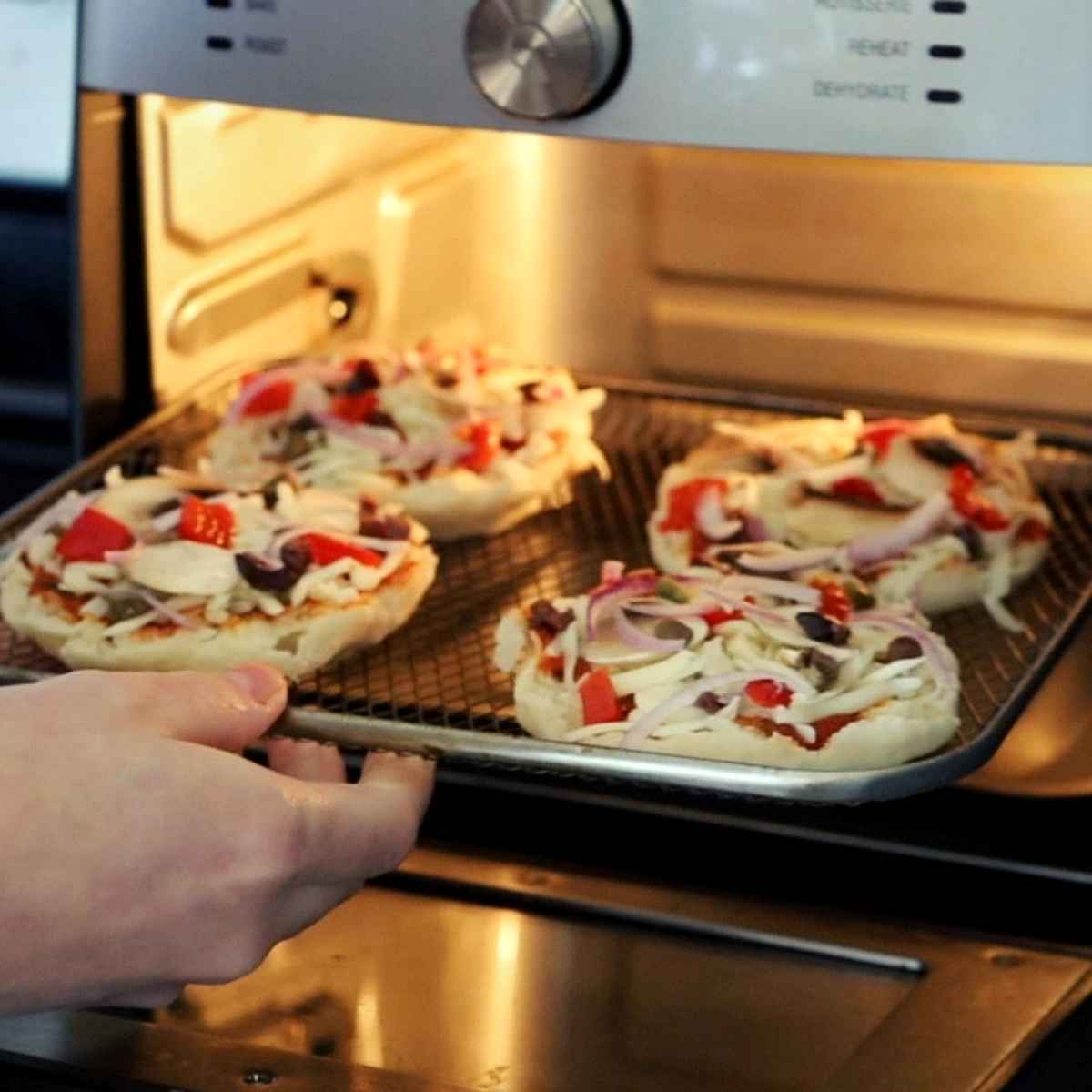 A hand sliding prepared english muffin pizzas into the air fryer for cooking.