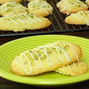 Freshly baked oval lime pistachio cookies topped with chopped pistachios and drizzled with white icing, displayed on a green plate with more cookies cooling on a wire rack in the background.