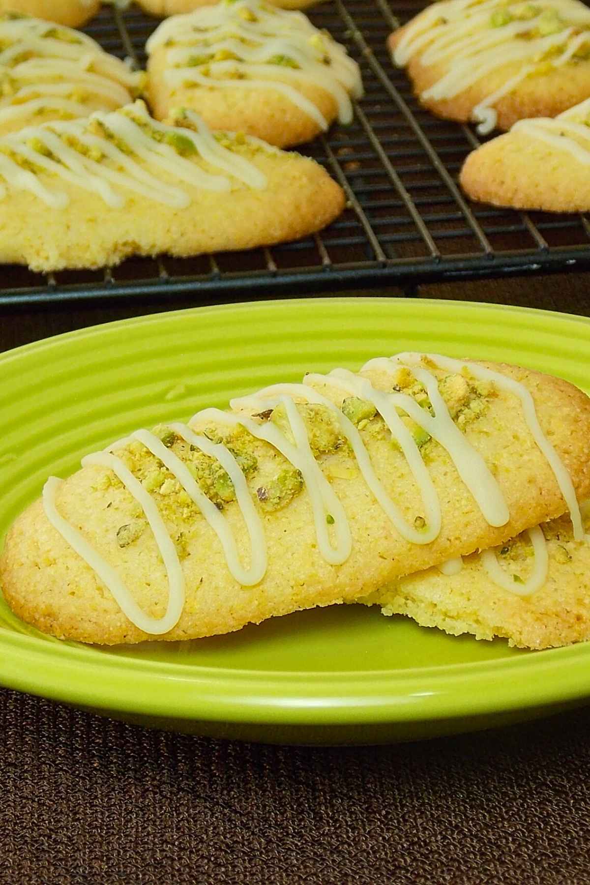 Freshly baked oval lime pistachio cookies topped with chopped pistachios and drizzled with white icing, displayed on a green plate with more cookies cooling on a wire rack in the background.