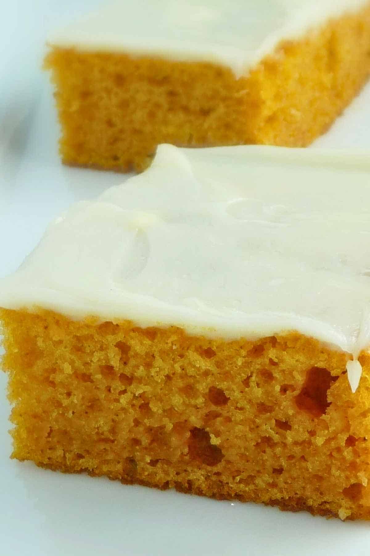 Upclose image of Two pieces of pumpkin sheet cake topped with cream. cheese icing on a white plate.