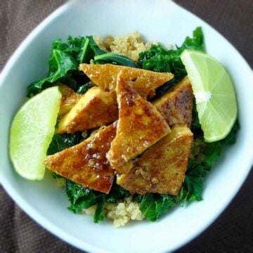 A white bowl filled with quinoa, steamed kale, and sweet chili lime tofu pieces garnished with two lime wedges.