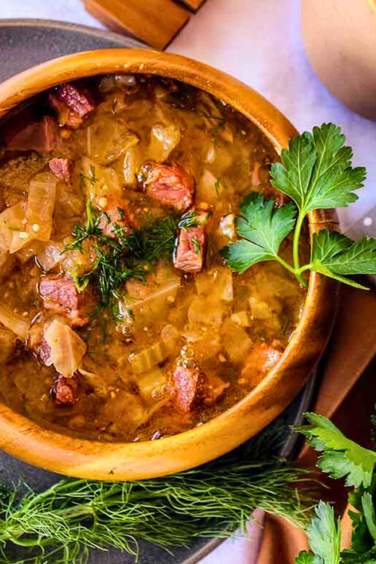 A top-down view of a rustic wooden bowl filled with hearty Corned Beef and Cabbage Soup, garnished with fresh fennel fronds and served with a sliced lemon on the side.
