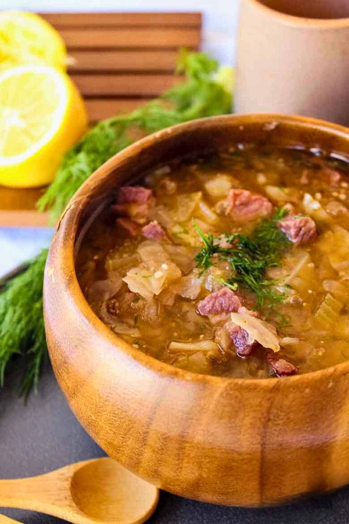 A top down view of a rustic wooden bowl filled with hearty corned beef and cabbage soup, garnished with fresh fennel fronds and served with a sliced lemon on the side.