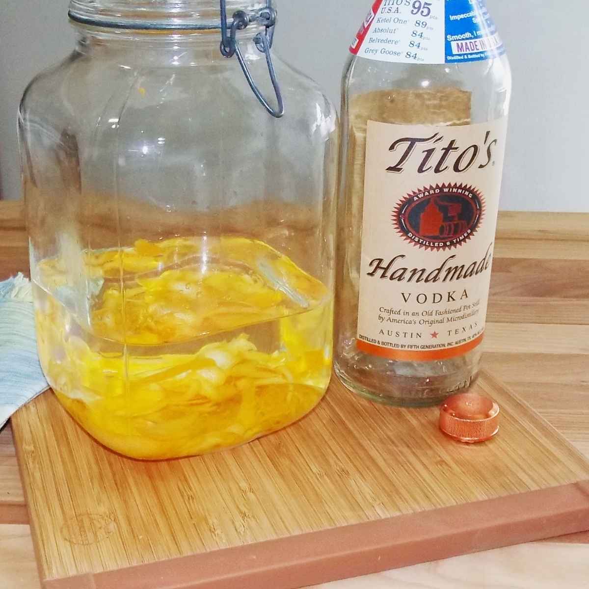 A clear glass gallon jar filled with lemon zest and vodka with a bottle of Tito's Handmade Vodka next to it.