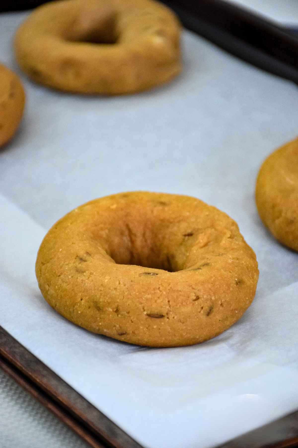 Unbaked bagel dough rings flecked with caraway seeds sitting on white parchment paper before baking.