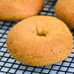 A close-up of a golden brown keto bagel resting on a black wire cooling rack, with two other bagels blurred in the background.