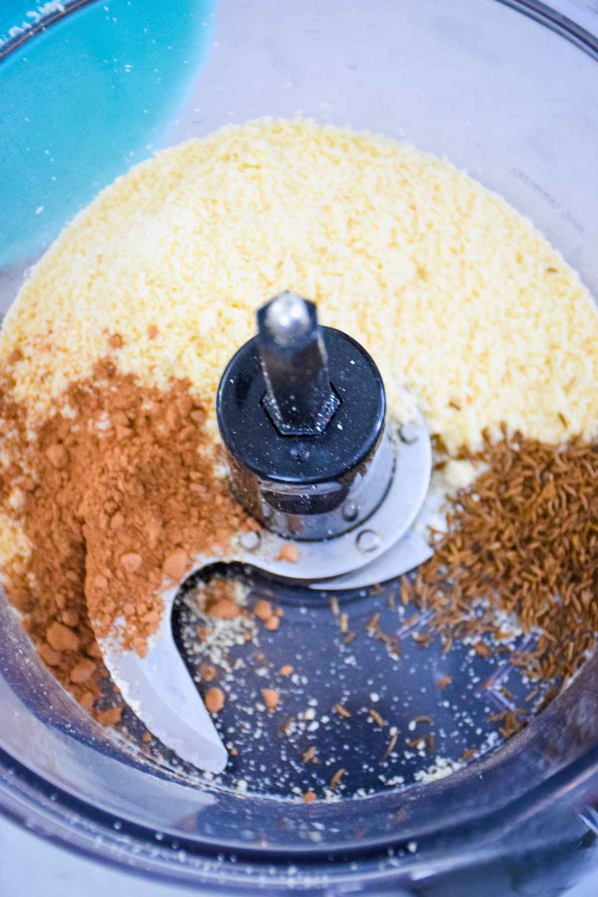 A top down view of a food processor bowl containing almond flour, caraway seeds, and cocoa powder, ready to be processed.