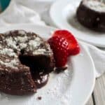 A close-up of a keto chocolate pudding cake with a molten center, topped with a dusting of powdered sweetener and served with a fresh strawberry slice on a white plate.