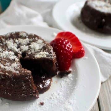 A close-up of a keto chocolate pudding cake with a molten center, topped with a dusting of powdered sweetener and served with a fresh strawberry slice on a white plate.