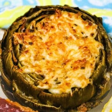 A close-up view of a keto stuffed artichoke featuring a bubbly, golden-brown melted cheese topping and savory herb filling.