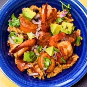 A deep blue bowl filled with a BBQ Shrimp Jambalaya containing seasoned rice, grilled shrimp, sliced sausage, and chunks of fresh avocado, red onion, and cilantro salad.