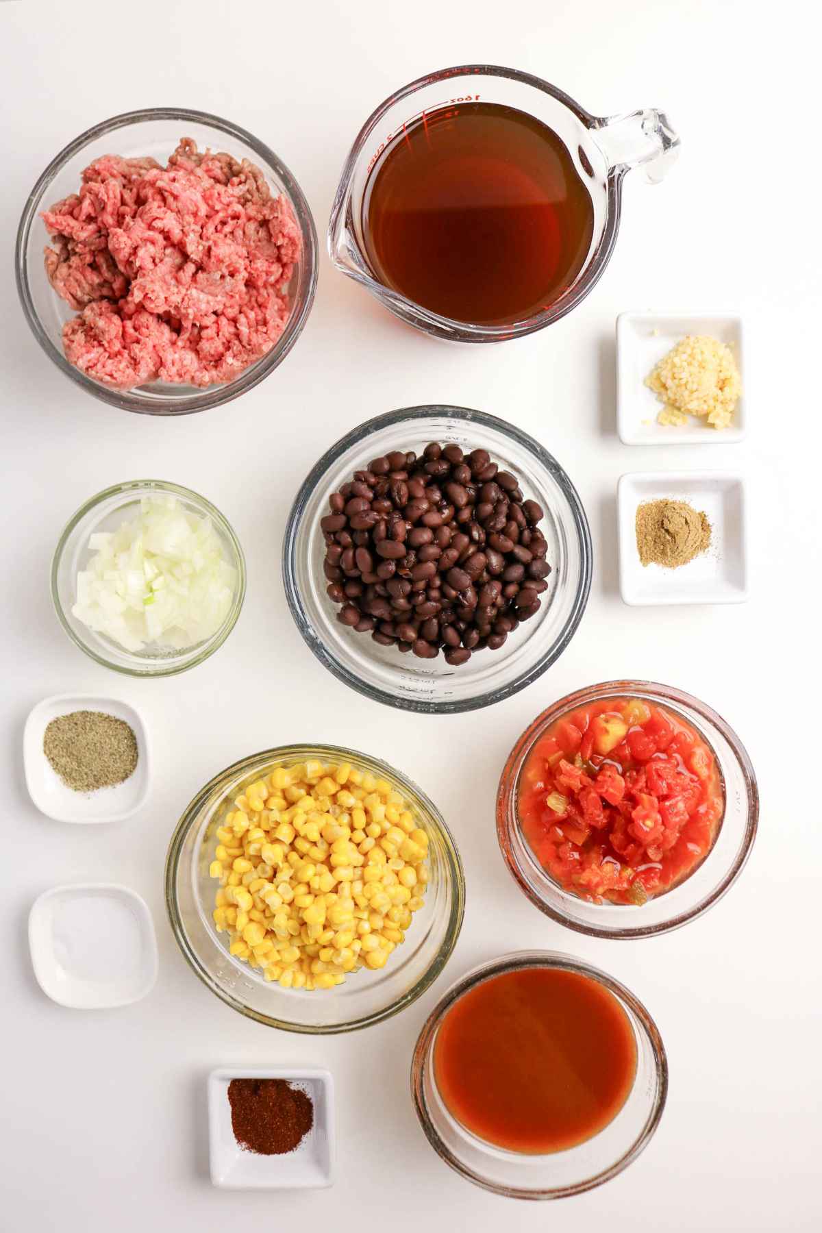 Flat lay of ingredients for soup in glass bowls including ground beef, beef broth, black beans, corn, diced tomatoes, enchilada sauce, onions, and various spices like chili powder and cumin.