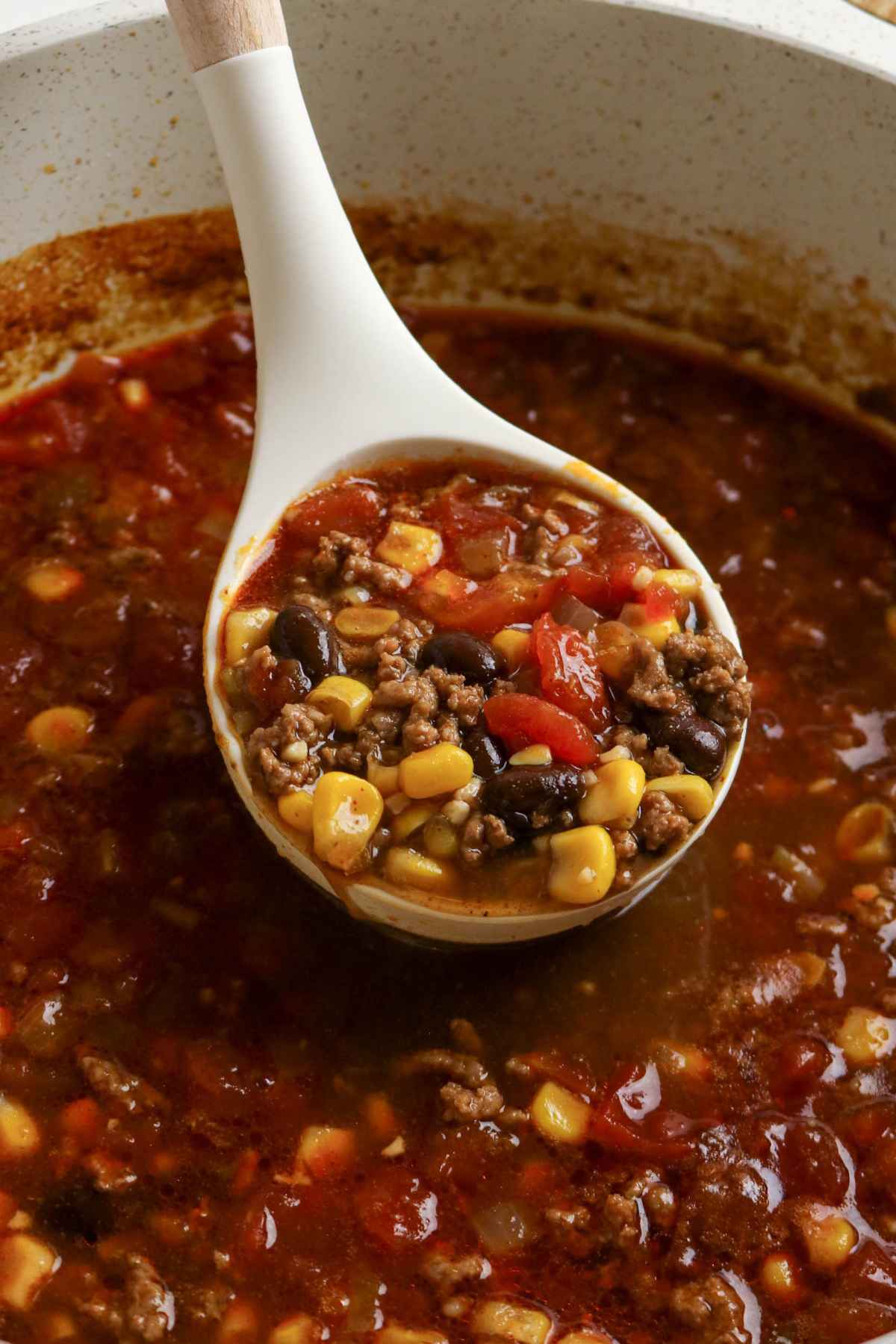 A ladle scooping a thick, hearty soup featuring ground meat, yellow corn, black beans, and tomatoes in a rich chili-based broth.