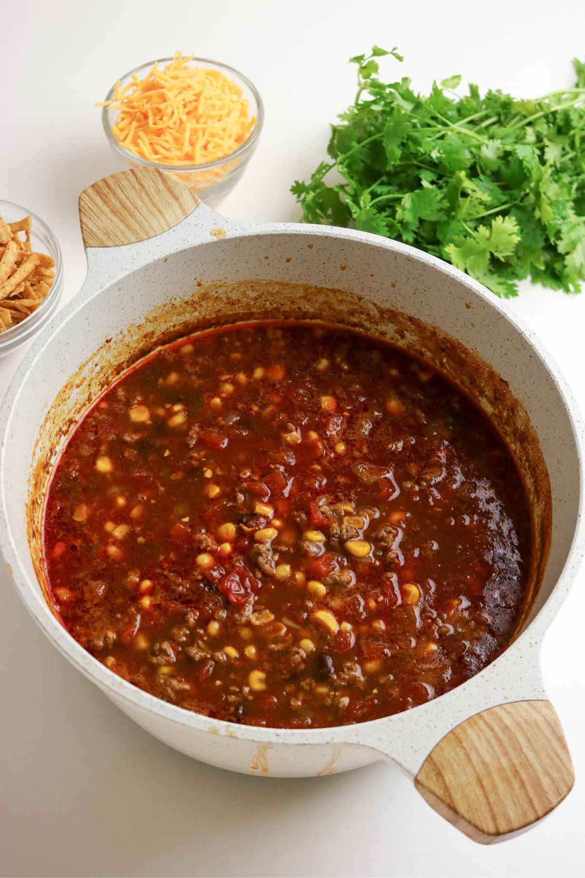 A white dutch oven filled with beef enchilada soup containing ground beef, corn, and beans, placed next to bowls of shredded cheese, tortilla strips, and fresh cilantro.
