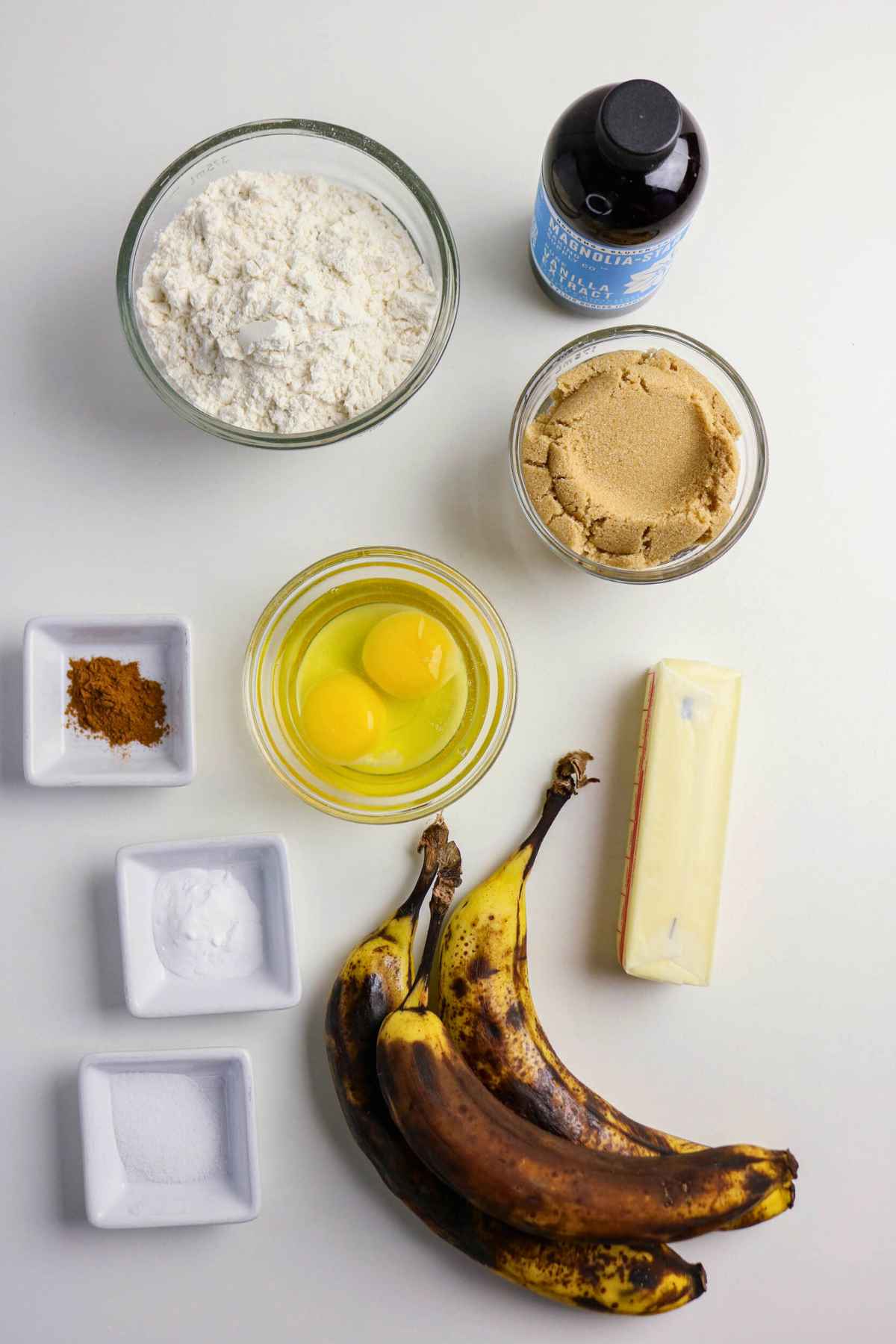 An overhead view of baking ingredients including overripe bananas, a stick of butter, two eggs in a glass bowl, flour, brown sugar, and small dishes of spices.