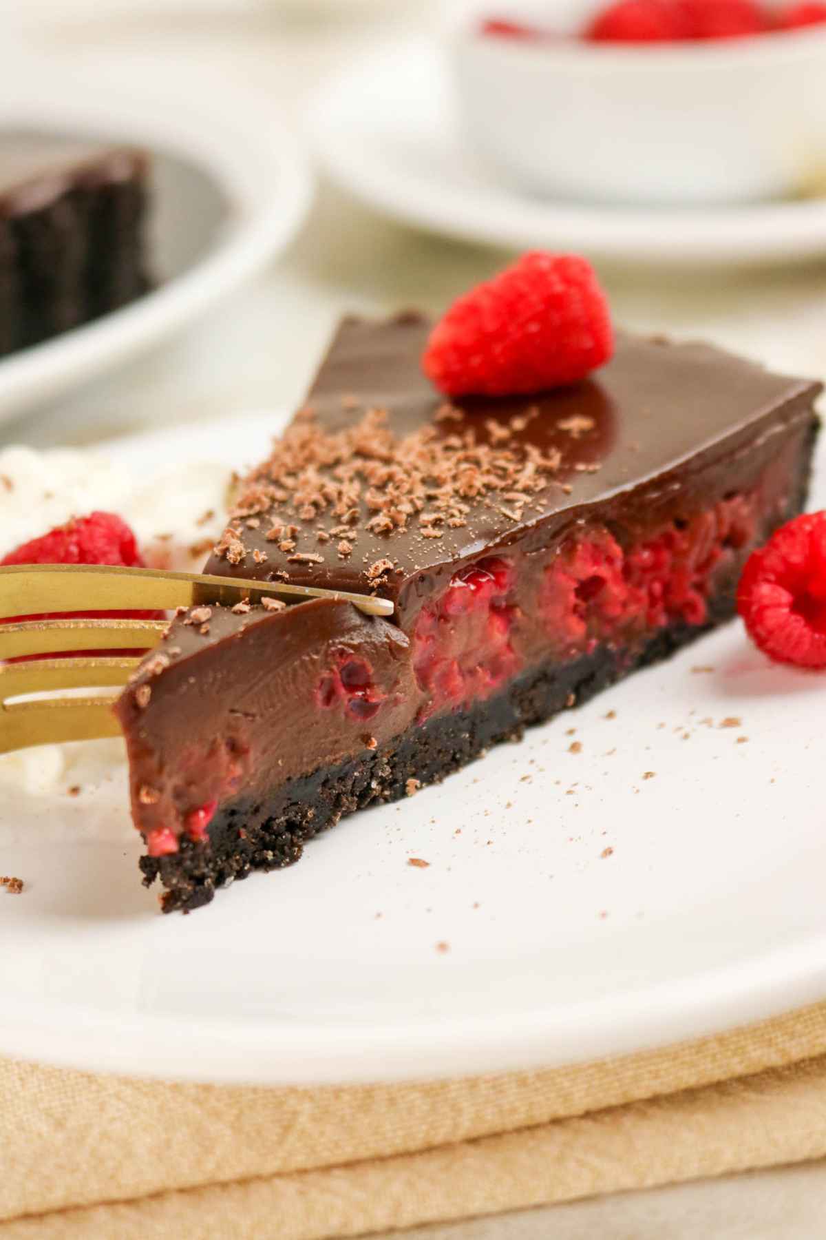 A close-up shot of a rich chocolate ganache tart slice on a white plate, showing the creamy chocolate filling, a layer of raspberries inside, and a gold fork cutting into the thart.