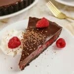 Slice of rich chocolate ganache raspberry tart on a white plate with a gold fork in the background.