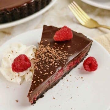 Slice of rich chocolate ganache raspberry tart on a white plate with a gold fork in the background.