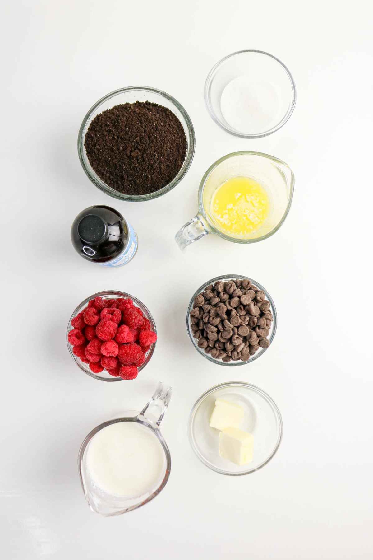 An overhead shot of recipe ingredients in glass bowls: chocolate cookie crust crumbs, fresh red raspberries, semi-sweet chocolate chips, heavy cream, melted butter, and vanilla extract.