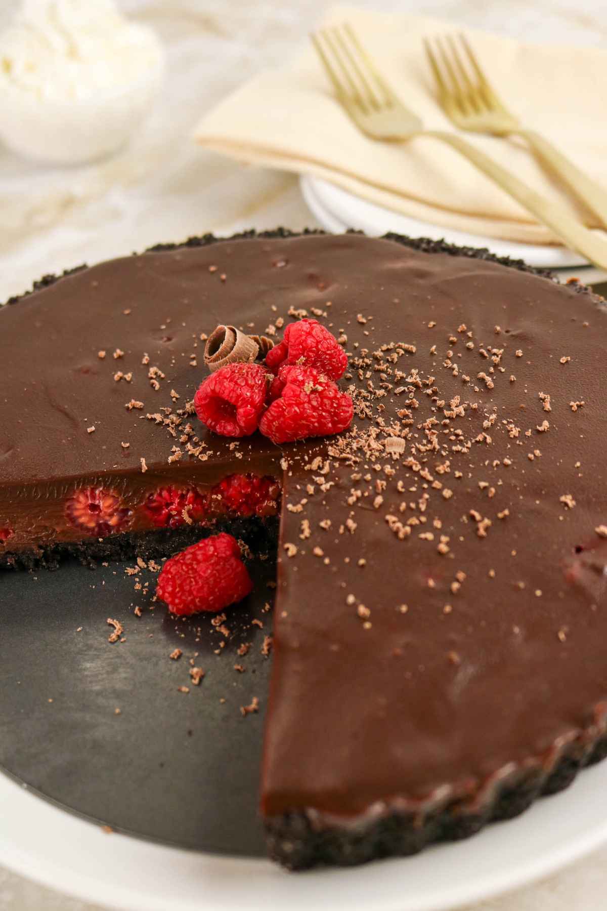 A whole chilled chocolate ganache tart with raspberries and a dark cookie crust, served on a white plate with gold forks in the background.
