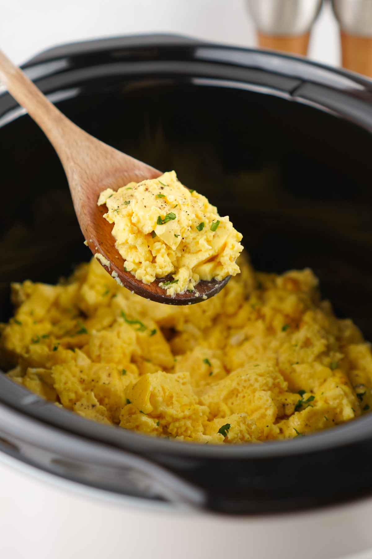 A wooden spoon scoops a serving of fluffy, eggs from a black slow cooker.