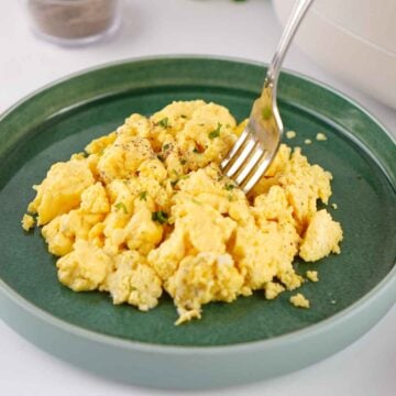 A silver fork takes a bite from a heap of fluffy, seasoned crockpot scrambled eggs served on a dark green plate.